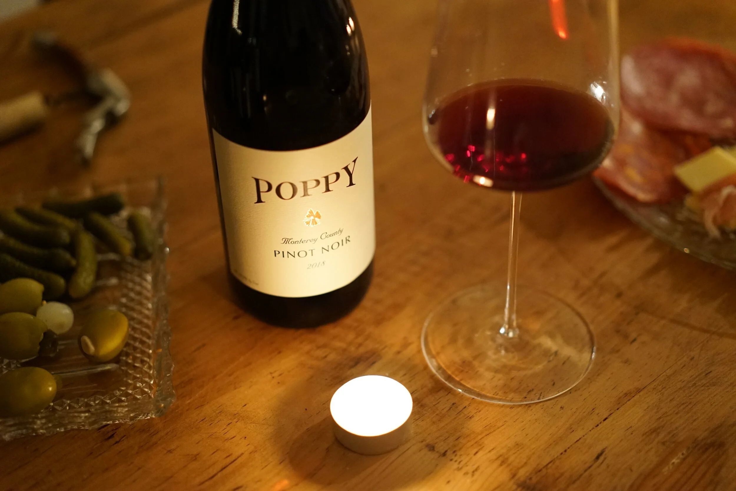 Poppy Pinot Noir bottle and glass with charcuterie and pickles on a wooden table.