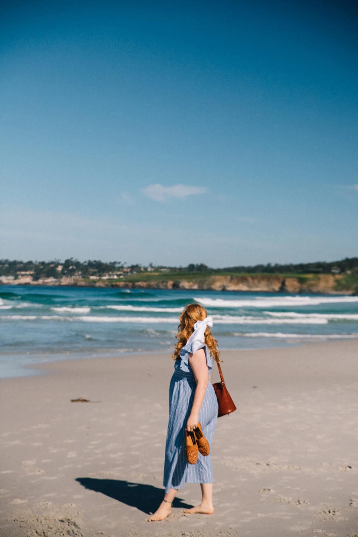 Woman with red hair in blue dress walks on beach, holding shoes.