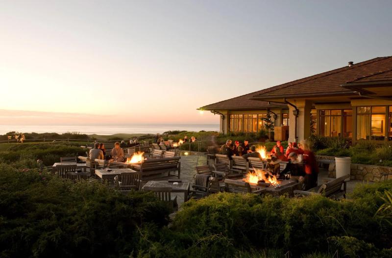 People gather around fire pits at a winery patio overlooking the ocean at sunset.