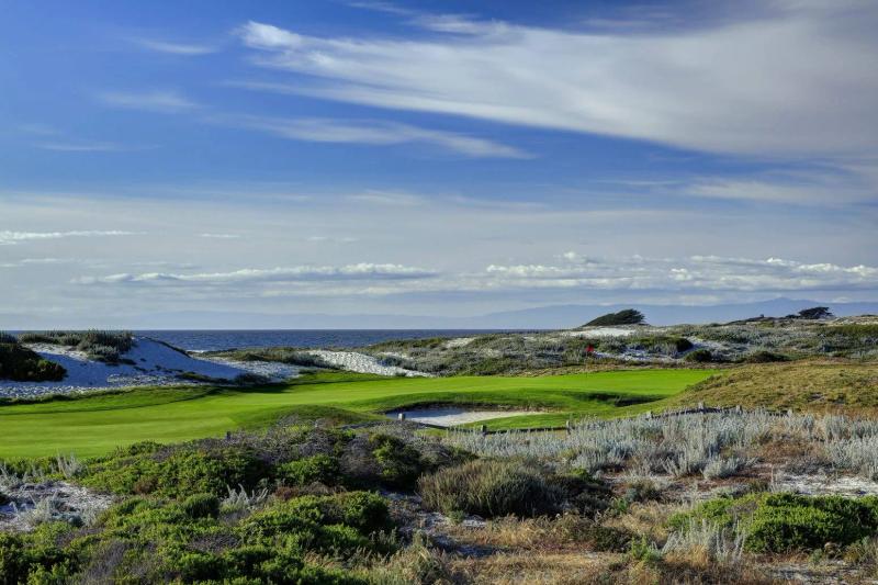 Coastal golf course with green fairways, sand dunes, and the Pacific Ocean under a blue sky.
