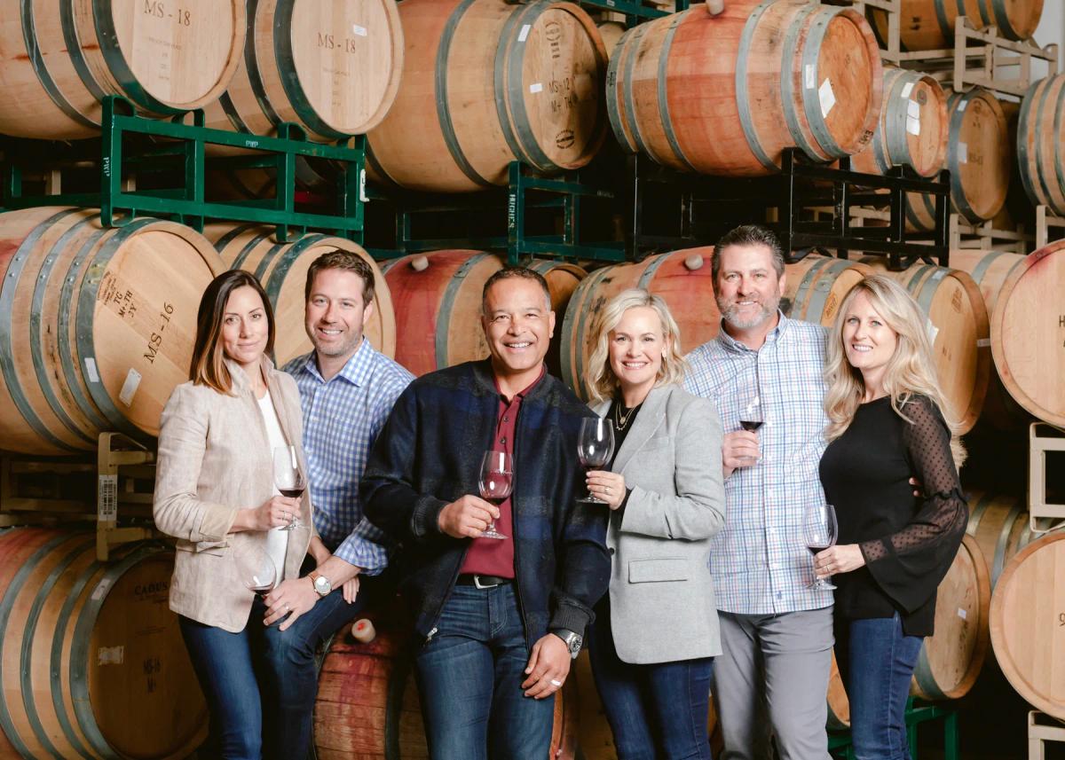 Six people holding glasses of red wine stand together in a winery barrel room, surrounded by rows of oak wine barrels stacked from floor to ceiling.