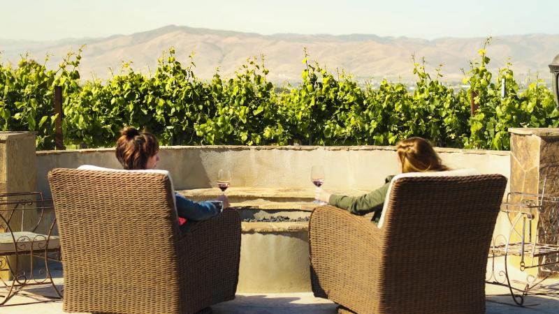Two people relax with wine by a fire pit at CRU Winery, overlooking Santa Lucia Highlands vineyards.