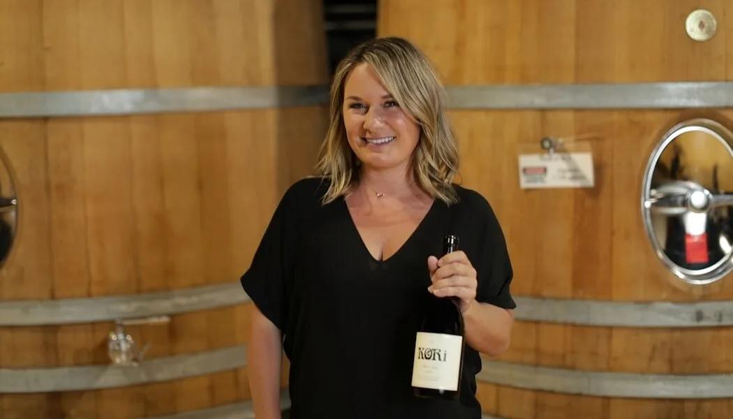 Kori Violini smiles holding a bottle of wine in front of large oak barrels in Santa Lucia Highlands.
