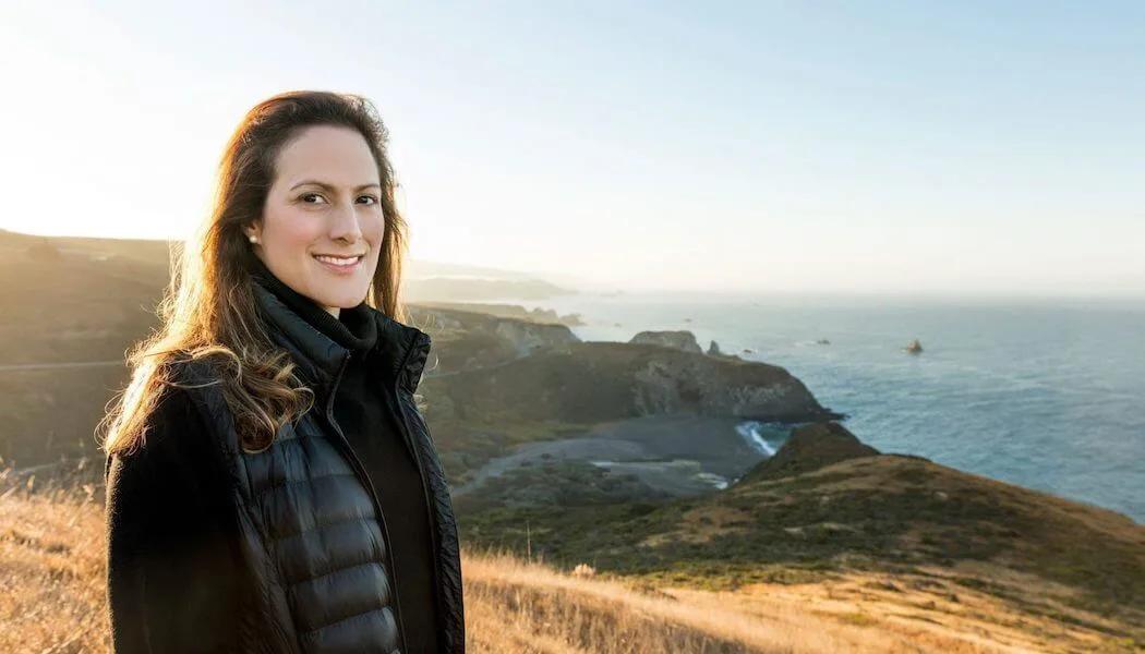 Woman in puffer vest smiles with coastal California hills and ocean behind her.